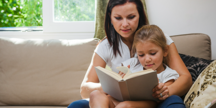 Mãe ensina filha a ler em casa durante momento de leitura no sofá.