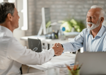 Dois homens adultos apertam as mãos sorridentes em mesa de escritório iluminada.