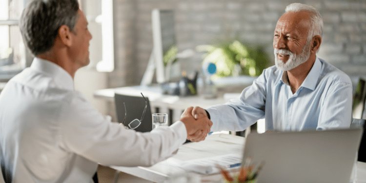 Dois homens adultos apertam as mãos sorridentes em mesa de escritório iluminada.