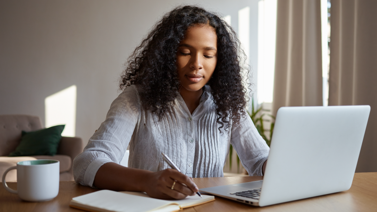 Jovem concentrada estudando em casa com caderno e notebook.