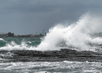 Ondas impactam rochas costeiras sob céu nublado em mar agitado.