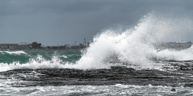Ondas impactam rochas costeiras sob céu nublado em mar agitado.