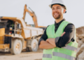Mestre de obras sorridente em obra com caminhão e escavadeira ao fundo.