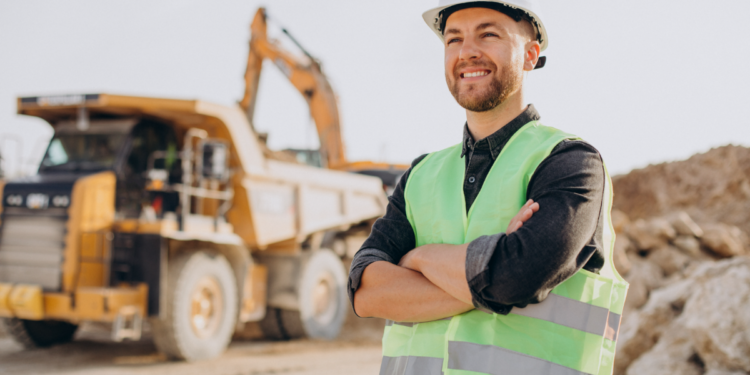 Mestre de obras sorridente em obra com caminhão e escavadeira ao fundo.