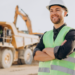 Mestre de obras sorridente em obra com caminhão e escavadeira ao fundo.