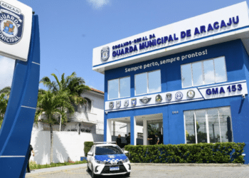 Fachada azul da Guarda Municipal de Aracaju com placa, viatura oficial e agentes na entrada e entorno.