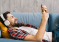 Homem ouvindo música relaxado no sofá com fone e celular.
