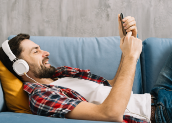 Homem ouvindo música relaxado no sofá com fone e celular.