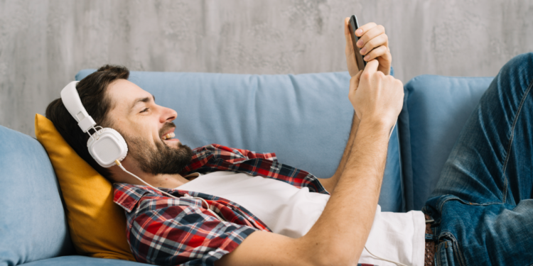 Homem ouvindo música relaxado no sofá com fone e celular.