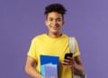 Jovem estudante sorrindo segura celular e cadernos em fundo roxo.