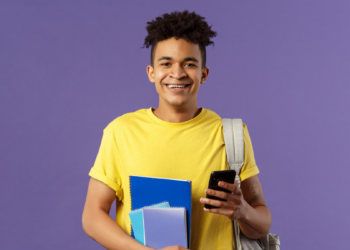 Jovem estudante sorrindo segura celular e cadernos em fundo roxo.