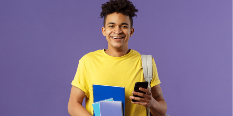 Jovem estudante sorrindo segura celular e cadernos em fundo roxo.