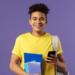 Jovem estudante sorrindo segura celular e cadernos em fundo roxo.