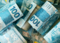 Cédulas de 100 reais brasileiras enroladas lado a lado em tom azul vibrante.