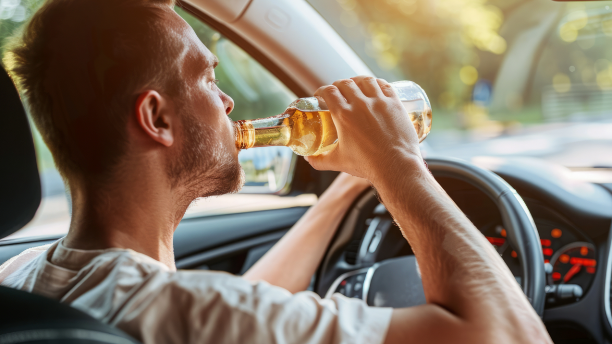 Jovem dirigindo enquanto consome bebida alcoólica em carro durante o dia.