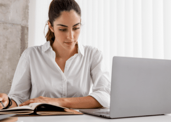 Jovem mulher concentrada estudando com notebook e caderno em ambiente claro.