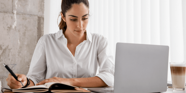 Jovem mulher concentrada estudando com notebook e caderno em ambiente claro.