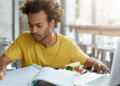 Jovem estudando concentrado com laptop, livros e lanche em cafeteria.