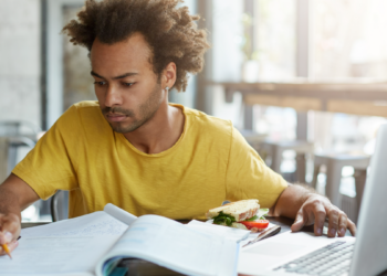 Jovem estudando concentrado com laptop, livros e lanche em cafeteria.