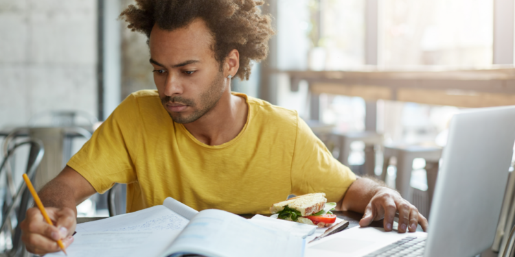 Jovem estudando concentrado com laptop, livros e lanche em cafeteria.