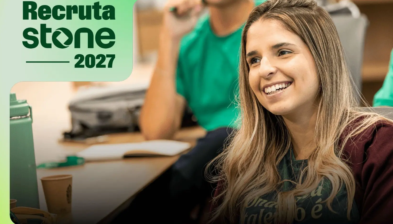 Mulher jovem de cabelos loiros sorrindo em sala com camiseta verde e logo Recruta Stone 2027 ao fundo.