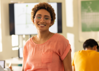 Professora sorridente em sala de aula moderna com estudantes ao fundo.