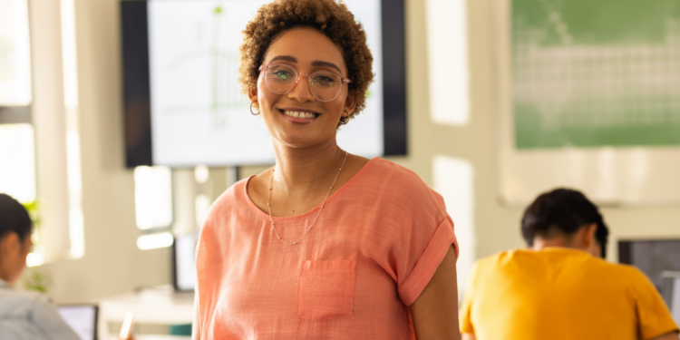 Professora sorridente em sala de aula moderna com estudantes ao fundo.