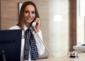 Recepcionista sorridente atende telefone em balcão de hotel.