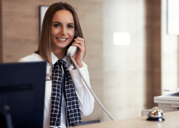 Recepcionista sorridente atende telefone em balcão de hotel.