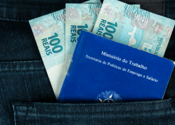 Carteira de trabalho azul ao lado de três notas de 100 reais em bolso de calça jeans.
