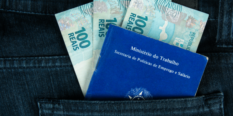 Carteira de trabalho azul ao lado de três notas de 100 reais em bolso de calça jeans.