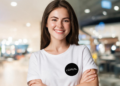 Jovem mulher de cabelo castanho liso com camiseta branca da L'Oréal sorrindo com braços cruzados em ambiente interno iluminado.