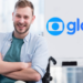 Jovem cadeirante sorridente em ambiente corporativo com logotipo da Globo ao fundo.