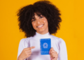 Jovem sorridente exibe carteira de trabalho brasileira em fundo amarelo.