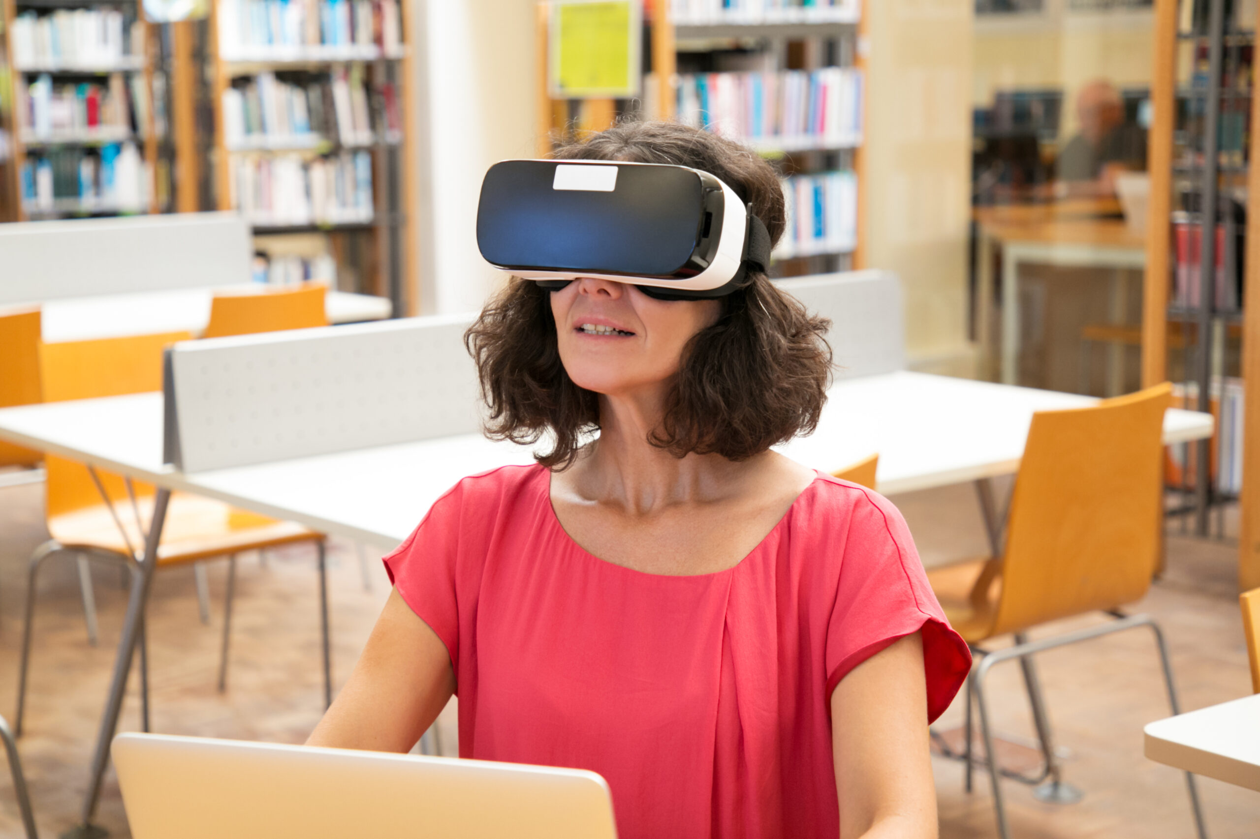 Mulher adulta usando óculos de realidade virtual em biblioteca moderna com mesas vazias, simbolizando os cursos gratuitos em IA.