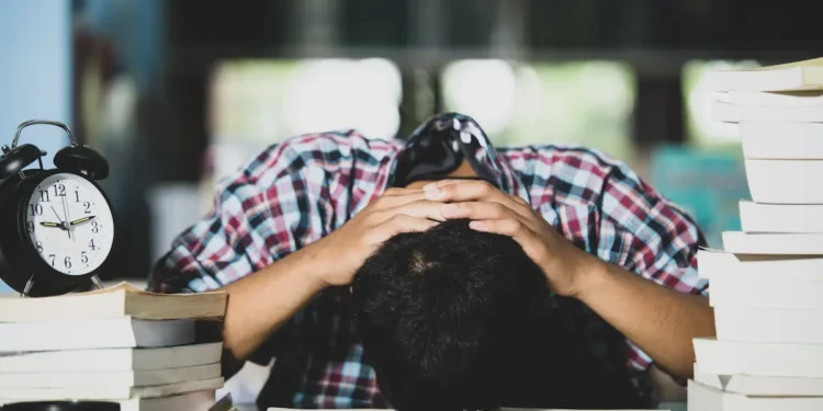 Estudante demonstra cansaço diante de livros e despertador durante uma rotina intensa de estudos.