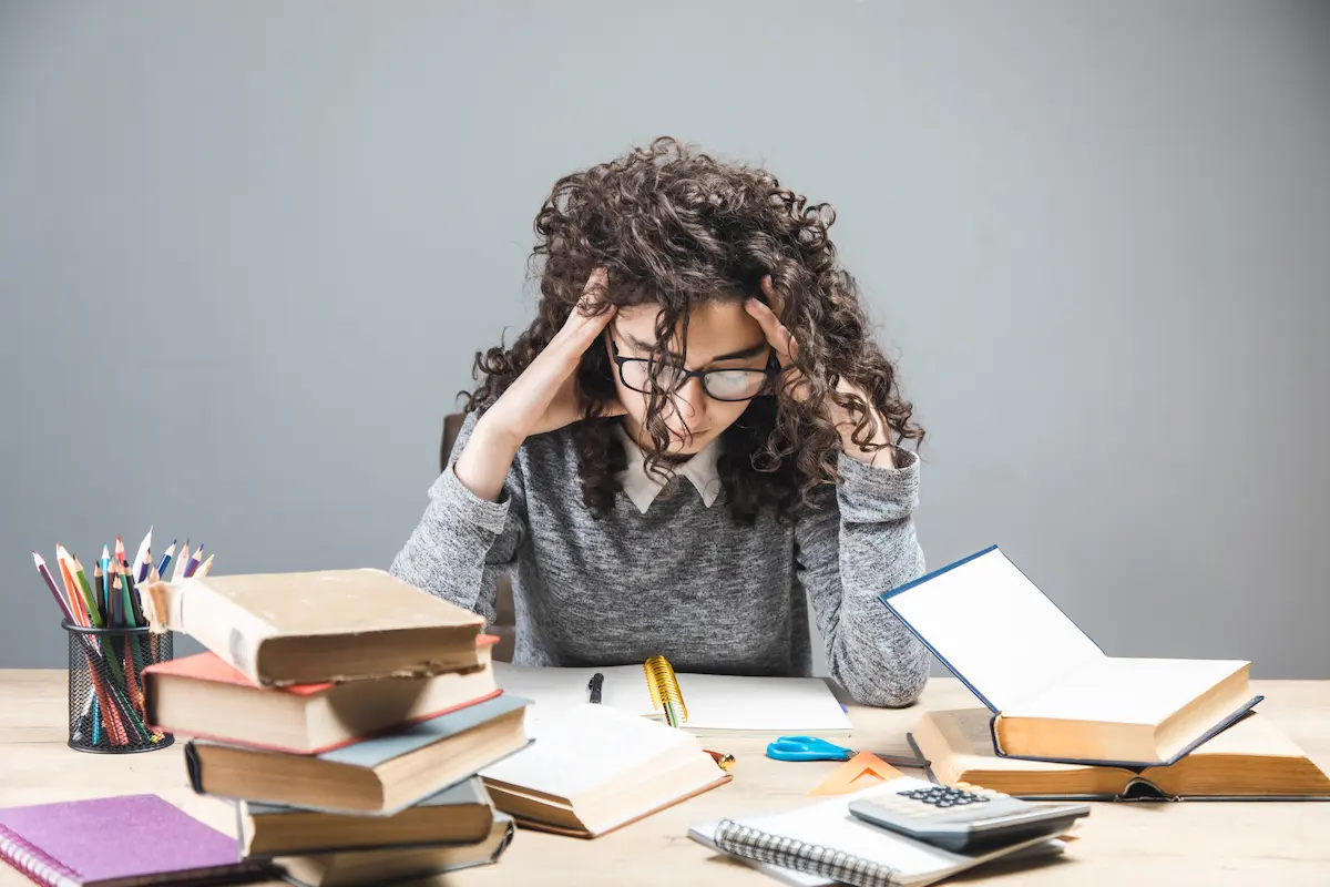 Estudante demonstra cansaço durante uma rotina intensa de estudos, cercada por livros e materiais escolares.