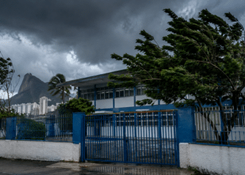 Fachada de escola pública no Rio de Janeiro com árvores curvadas pelo vento e céu escuro de tempestade, com o Cristo Redentor ao fundo, referência às unidades de ensino afetadas pela suspensão das aulas