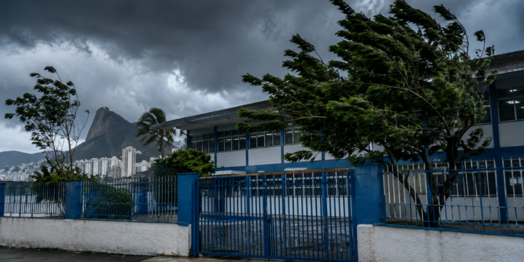 Fachada de escola pública no Rio de Janeiro com árvores curvadas pelo vento e céu escuro de tempestade, com o Cristo Redentor ao fundo, referência às unidades de ensino afetadas pela suspensão das aulas