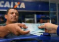 Trabalhador recebe cédulas de R$ 100 em atendimento bancário; salário mínimo pode chegar a R$ 1.741 em 2027.