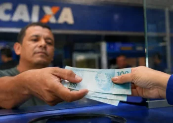 Trabalhador recebe cédulas de R$ 100 em atendimento bancário; salário mínimo pode chegar a R$ 1.741 em 2027.