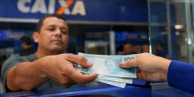 Trabalhador recebe cédulas de R$ 100 em atendimento bancário; salário mínimo pode chegar a R$ 1.741 em 2027.