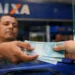 Trabalhador recebe cédulas de R$ 100 em atendimento bancário; salário mínimo pode chegar a R$ 1.741 em 2027.