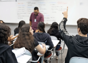 Foto de sala de aula com estudantes sentados em carteiras, um deles com a mão levantada, e o professor em pé junto ao quadro branco, remetendo ao ensino nos institutos federais alcançados pela autorização de vagas