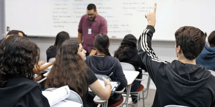 Foto de sala de aula com estudantes sentados em carteiras, um deles com a mão levantada, e o professor em pé junto ao quadro branco, remetendo ao ensino nos institutos federais alcançados pela autorização de vagas