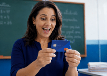 Imagem ilustrativa de professora sorrindo surpresa ao segurar um cartão azul diante de um quadro verde com contas de matemática, referência aos benefícios financeiros oferecidos aos docentes