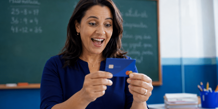 Imagem ilustrativa de professora sorrindo surpresa ao segurar um cartão azul diante de um quadro verde com contas de matemática, referência aos benefícios financeiros oferecidos aos docentes