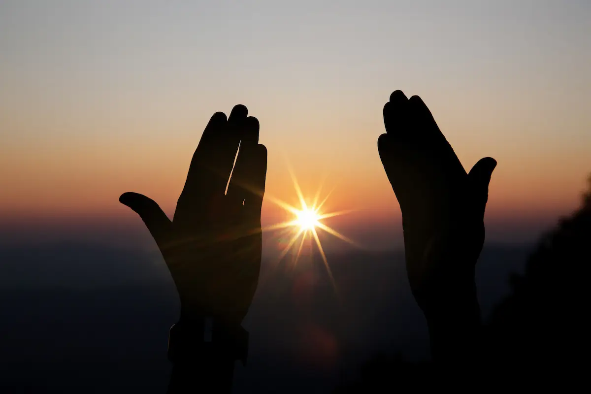Mãos erguidas diante do pôr do sol, simbolizando bênção, fé e espiritualidade.