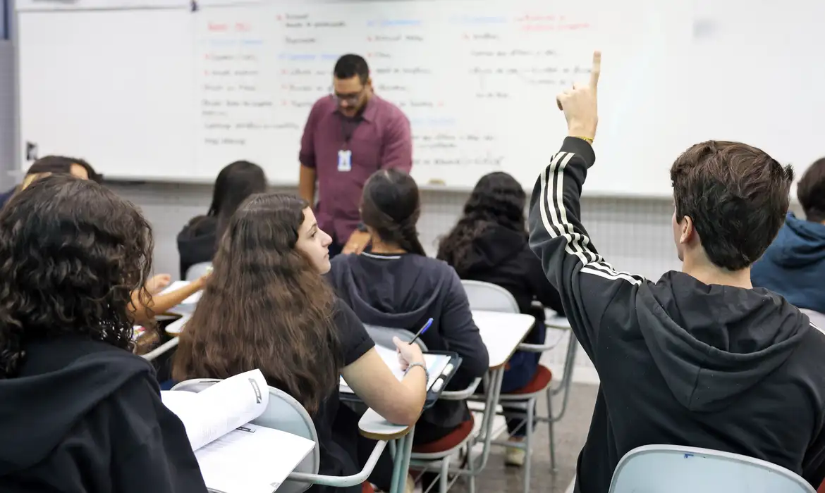 Foto de sala de aula com estudantes sentados em carteiras, um deles com a mão erguida, e o professor em pé junto ao quadro branco, referência ao público docente contemplado pelo programa federal