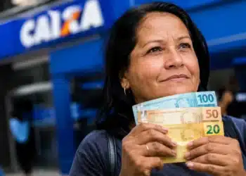 Mulher segura cédulas de real em frente a uma agência da Caixa, representando o período de pagamentos dos benefícios do INSS de até um salário mínimo em agosto.